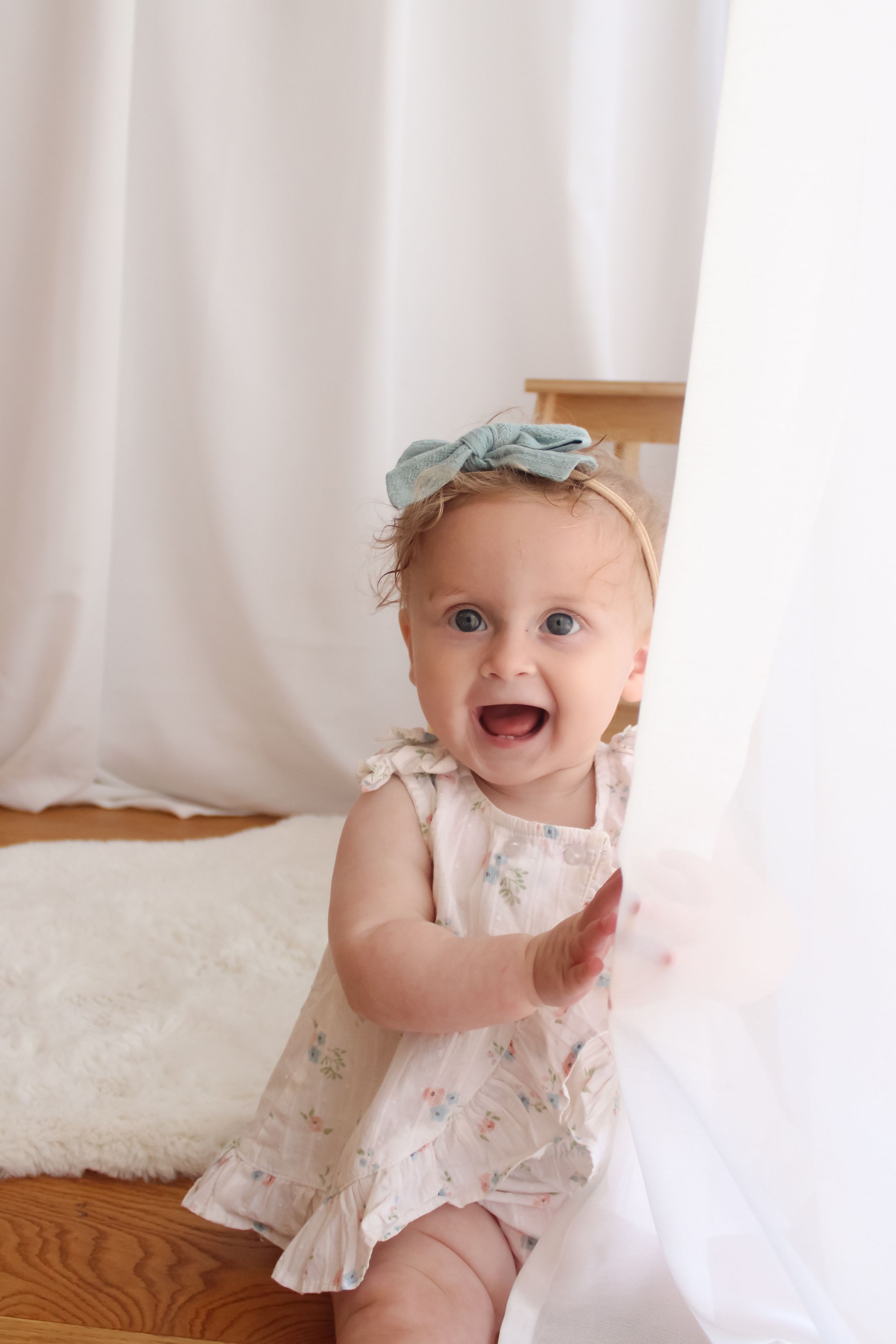 Baby in a white floral dress and mint headband plays with sheer curtains at a window during a photo shoot.