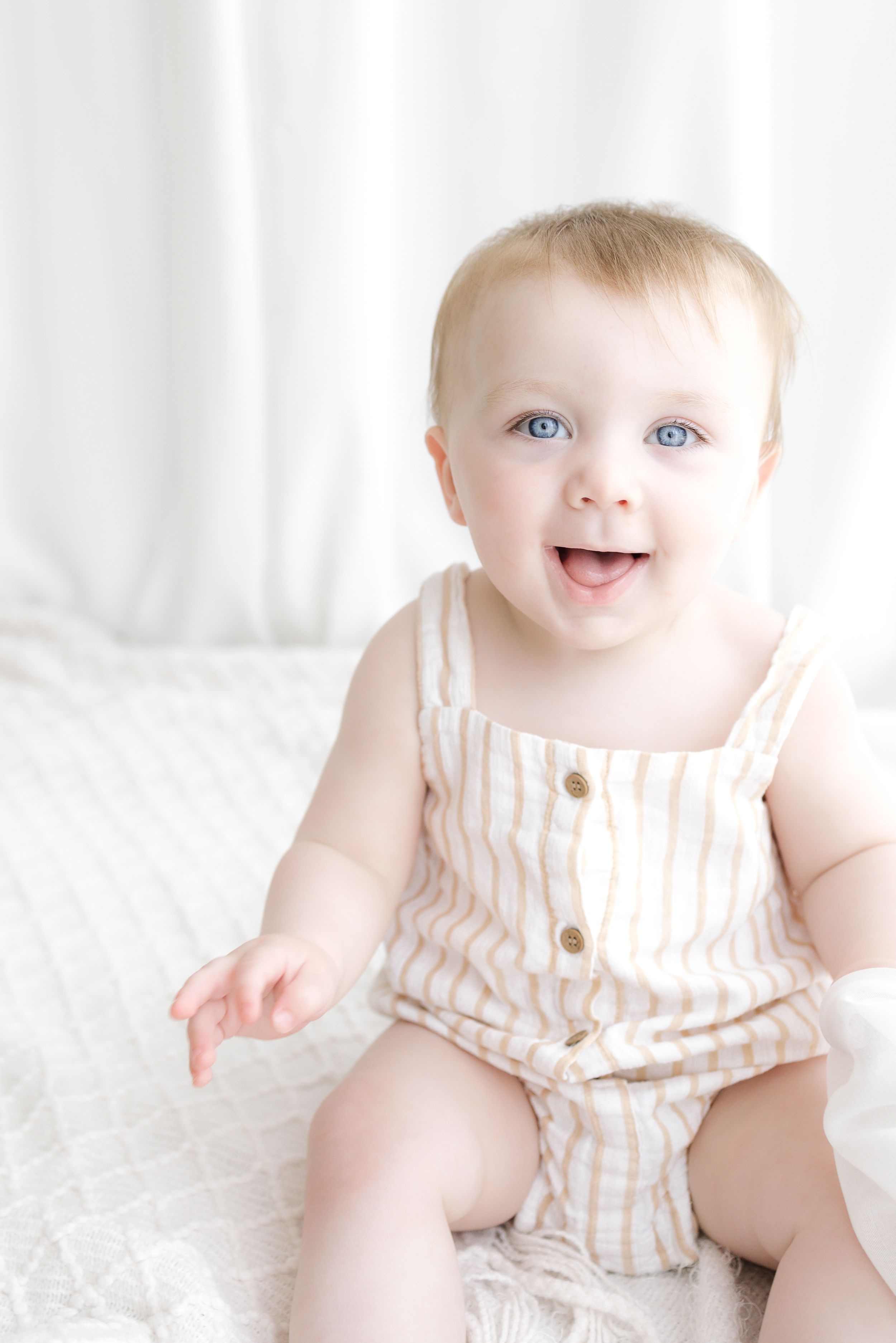 Happy baby with blue eyes sitting on white bed wearing striped romper, smiling joyfully in bright natural light.