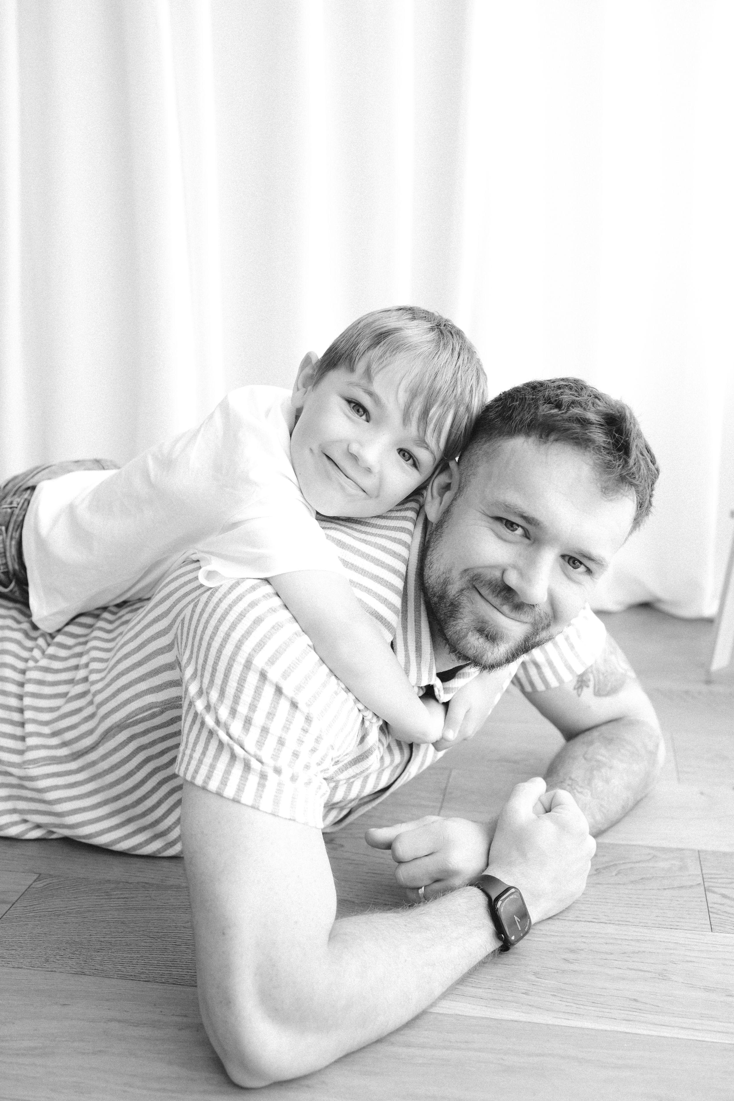 Black and white photo of a smiling father lying on floor with happy young son on his back.