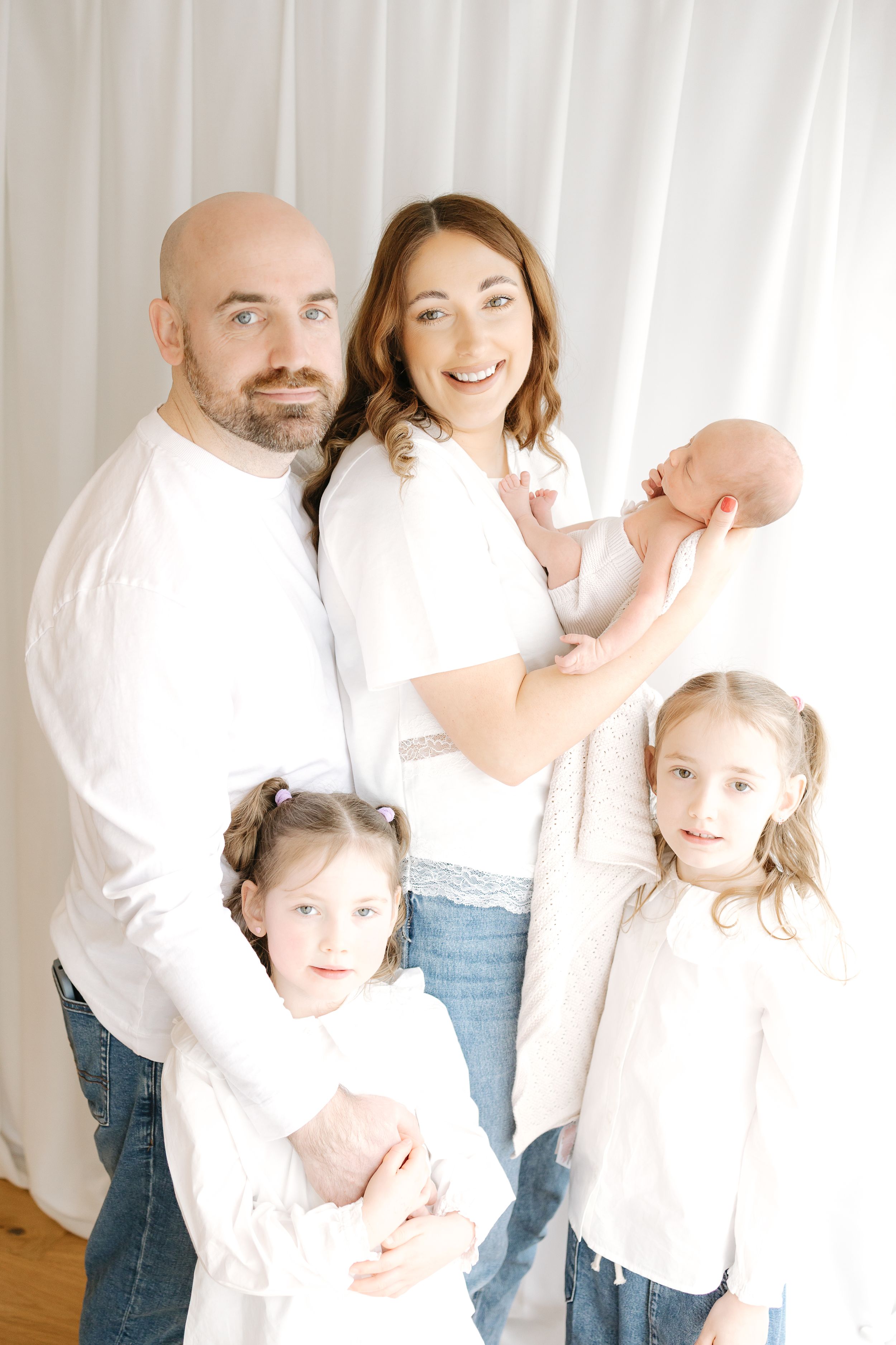A happy family of five in white tops and jeans, parents with three daughters including a newborn baby, posing for a portrait.