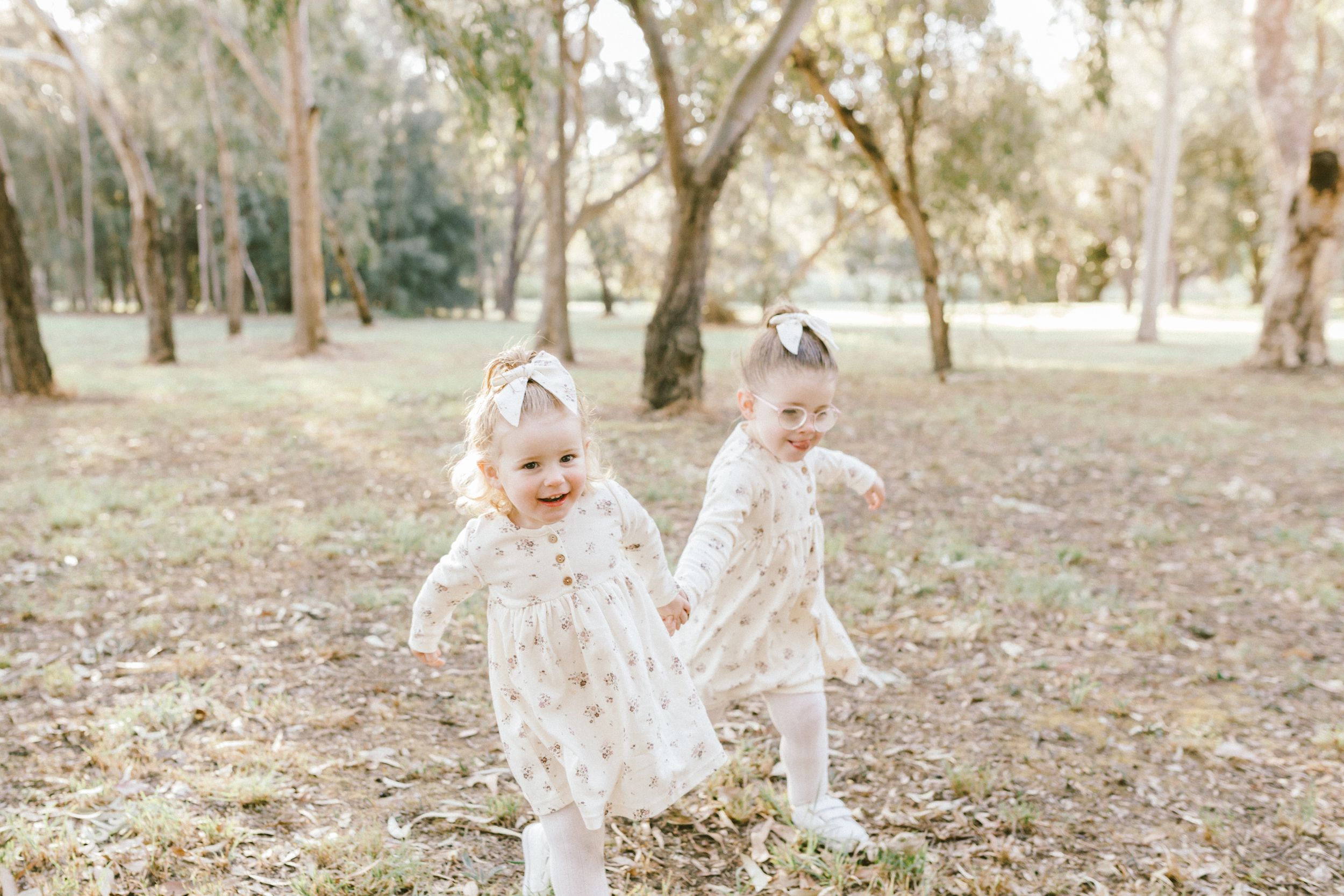 Young siblings dressed in white outfits play together on fallen autumn leaves in a wooded outdoor setting.