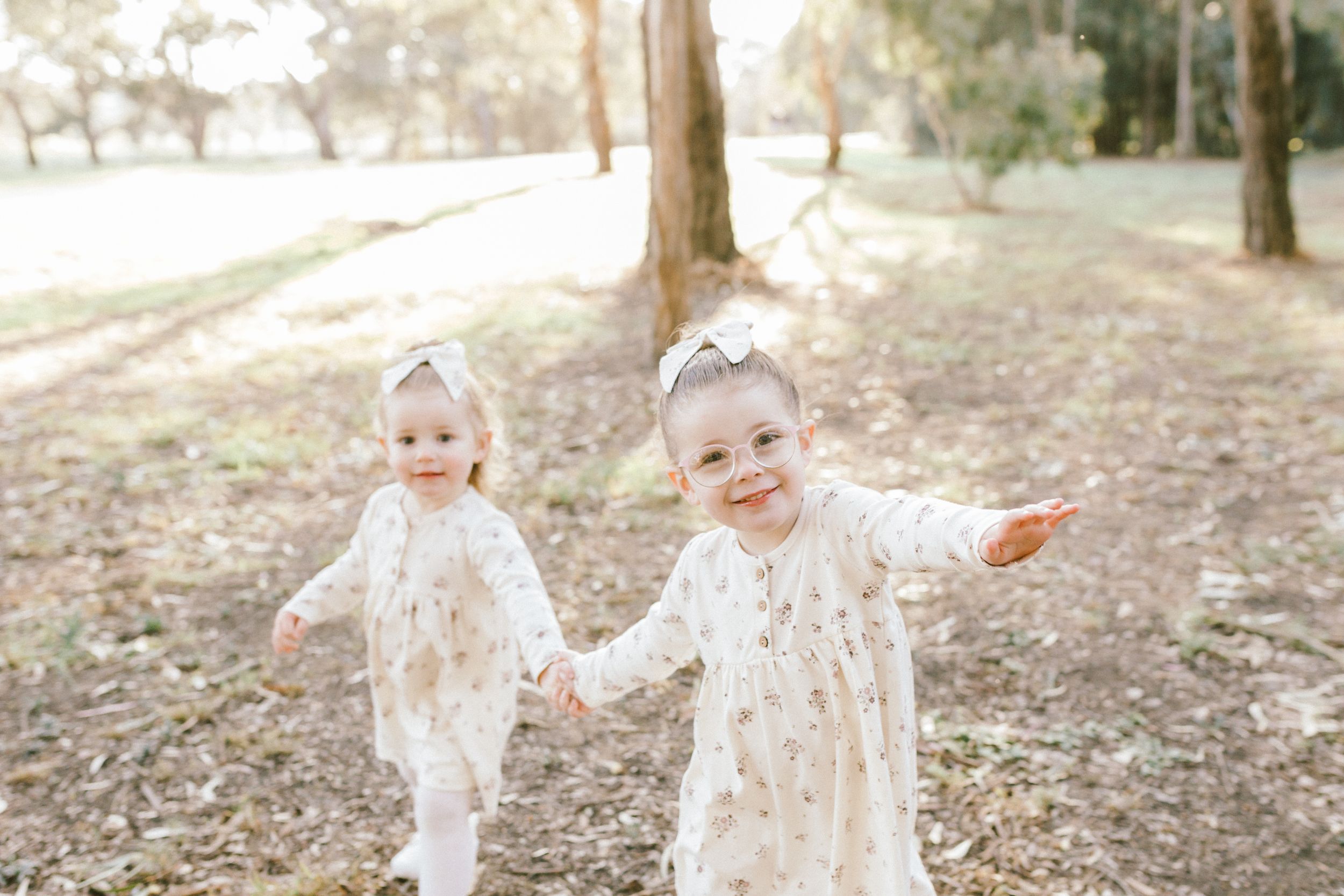 Sweet toddler in a white lace dress plays and twirls joyfully in a sunlit forest setting with autumn leaves scattered around.