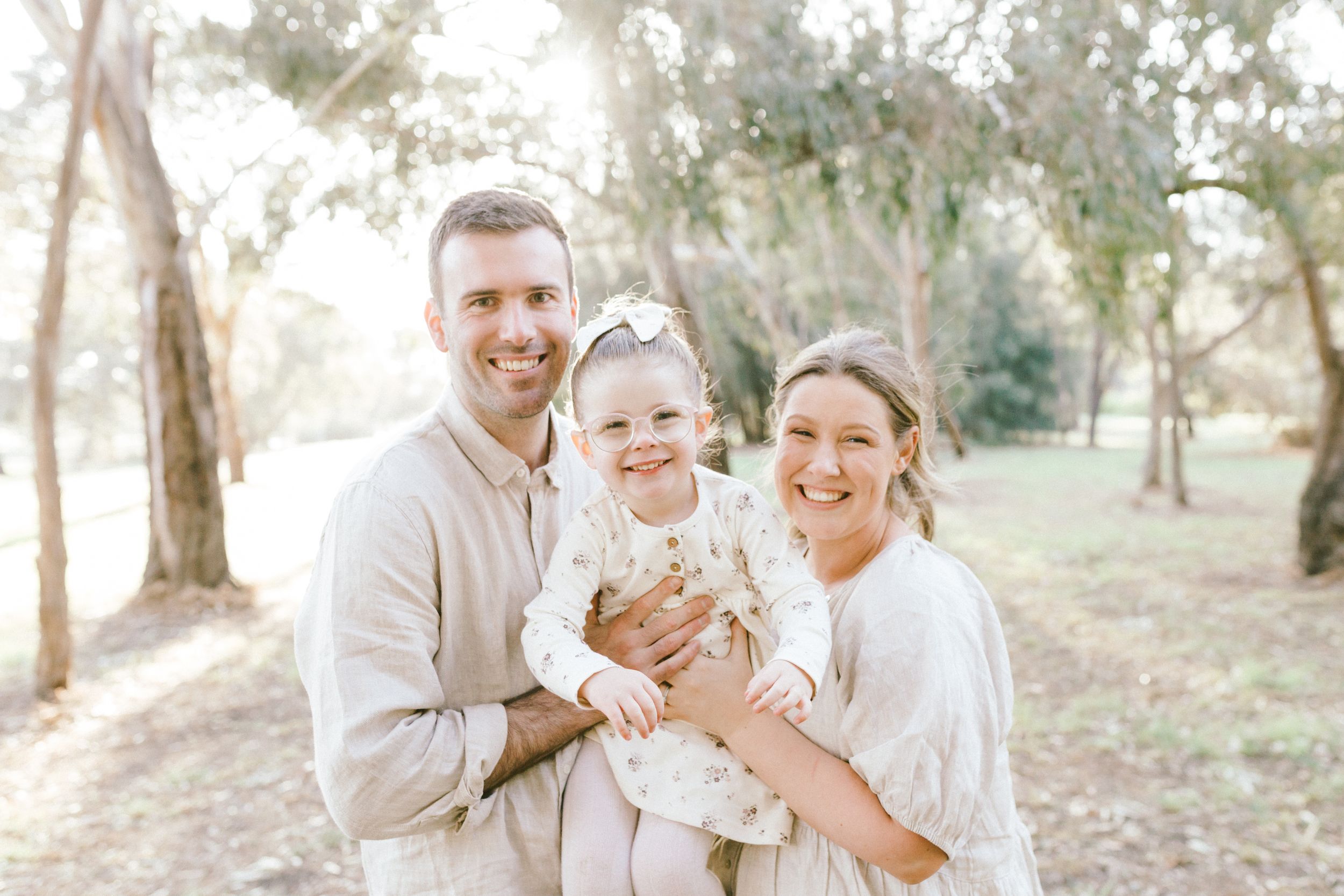 A family wearing coordinated beige and white outfits poses together in a sun-dappled forest setting.