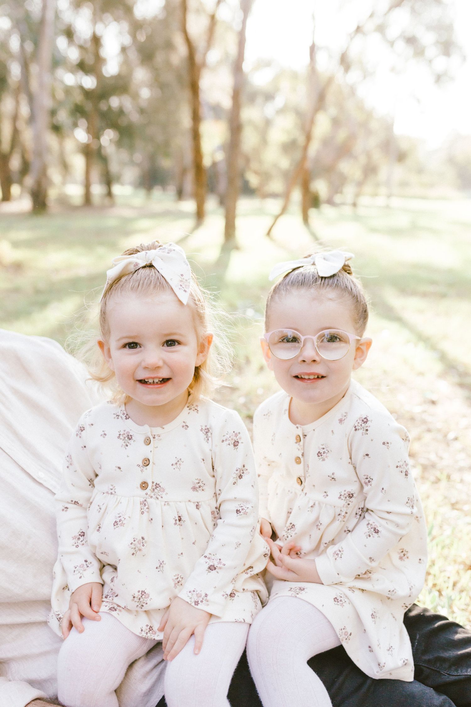 Two toddlers wearing matching floral outfits sit together and smile during an outdoor photoshoot in natural sunlight.