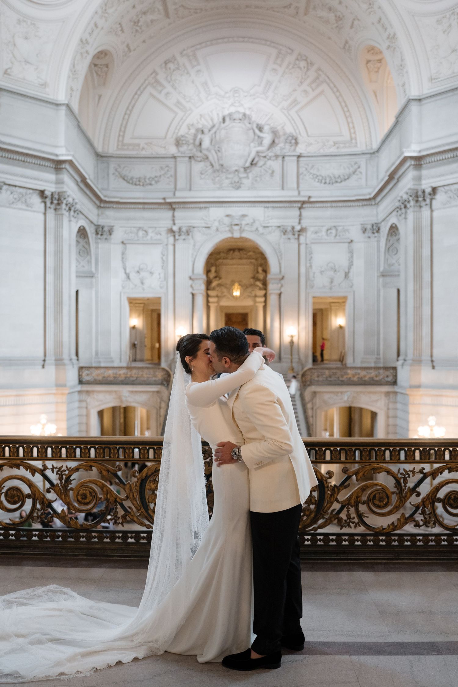 A romantic couple in wedding attire embraces in an elegant white marble hallway with ornate architectural details.
