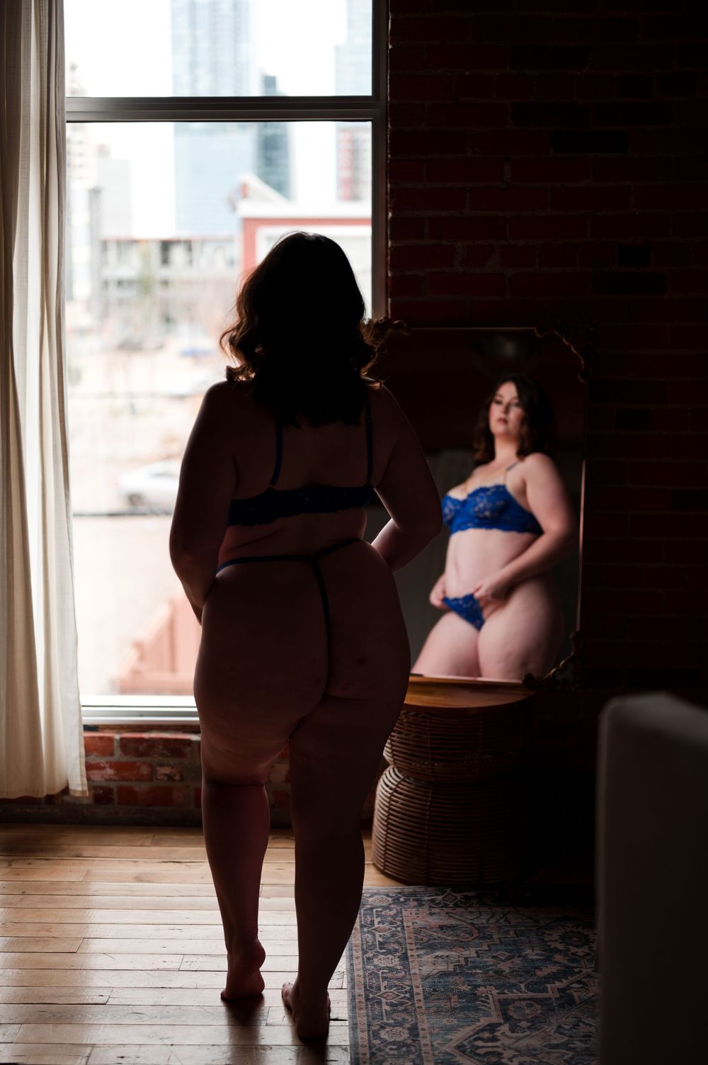Figure in silhouette stands before a mirror wearing blue lingerie in a sunlit room with exposed brick walls.