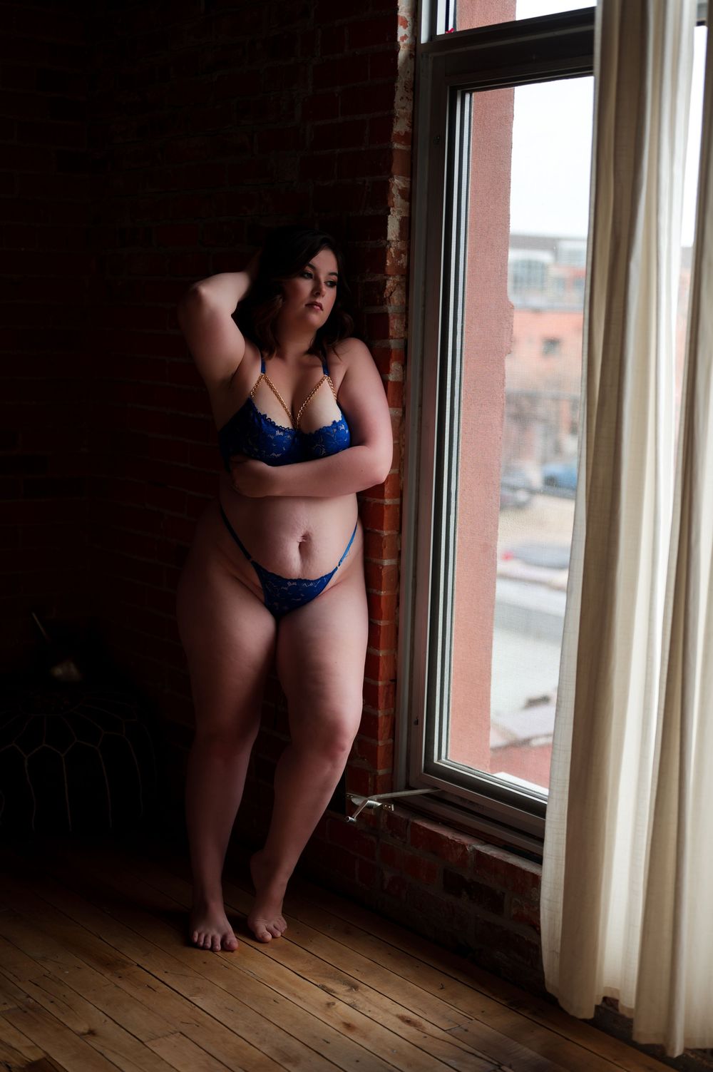 A person in blue lingerie poses in front of a window with white curtains on wooden floors in dramatic lighting.