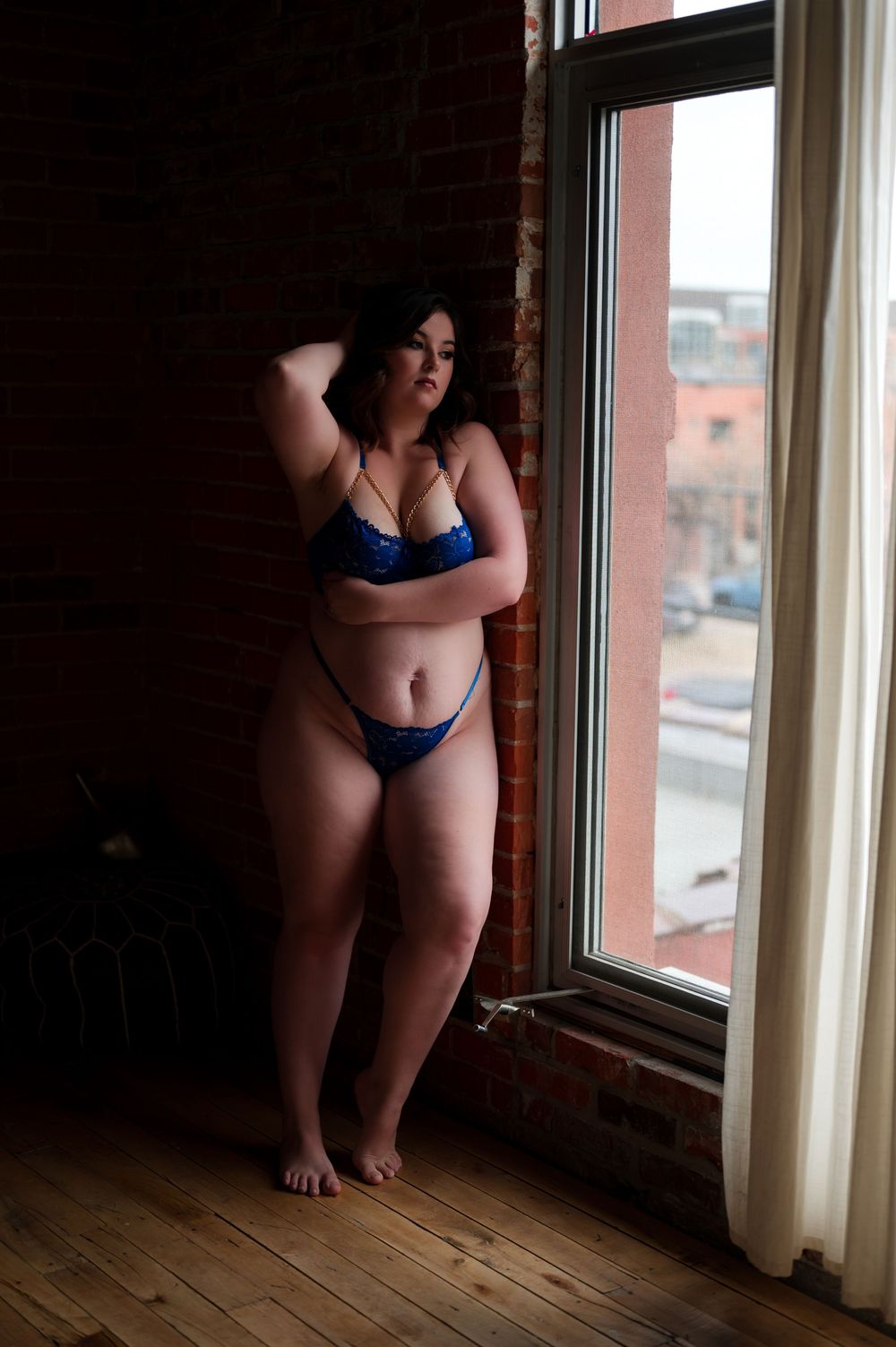 Person poses in blue lingerie by a window in an urban loft with wooden floors and exposed brick.