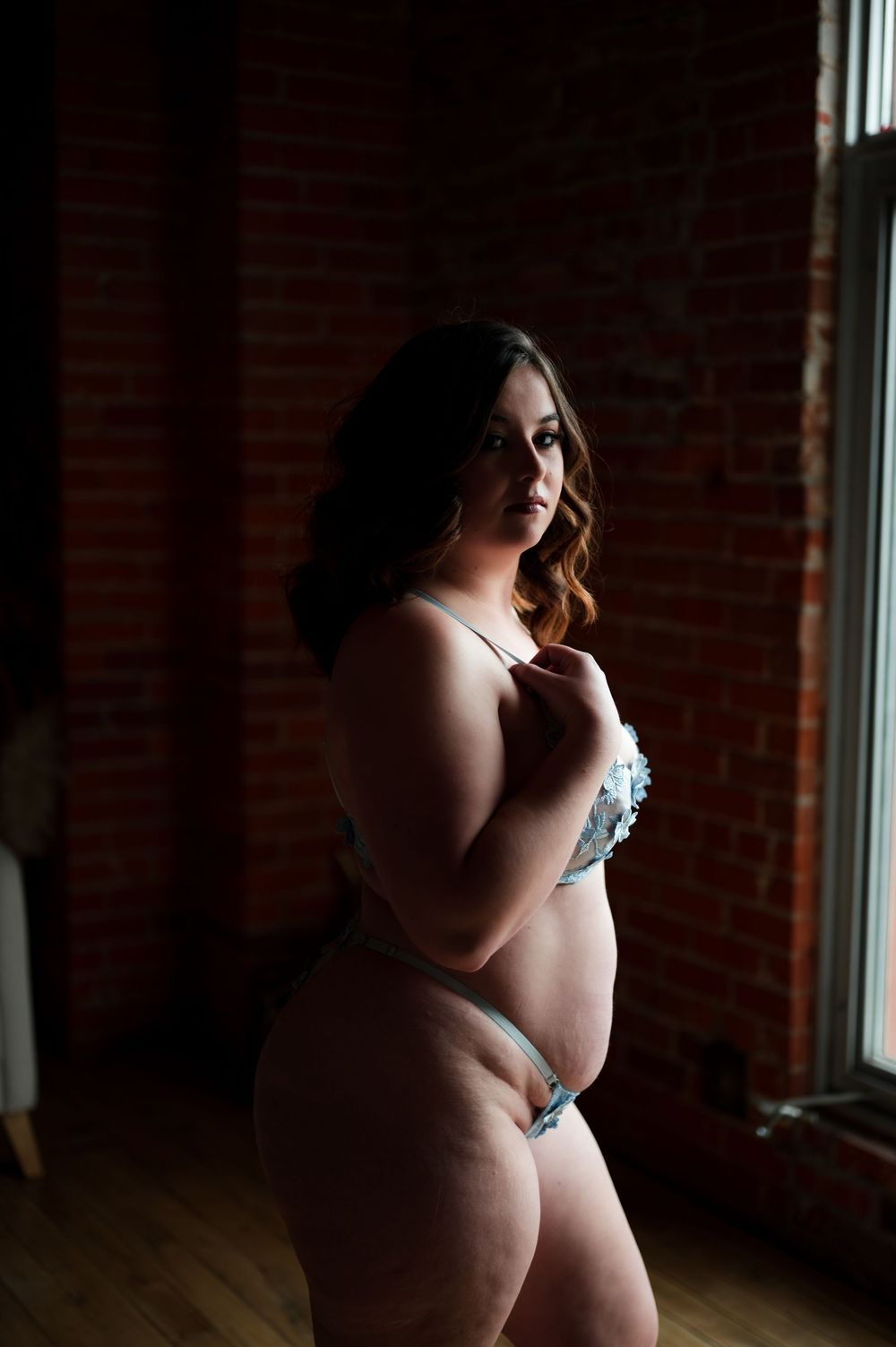 Artistic boudoir portrait in blue lingerie against exposed brick wall with dramatic window lighting.