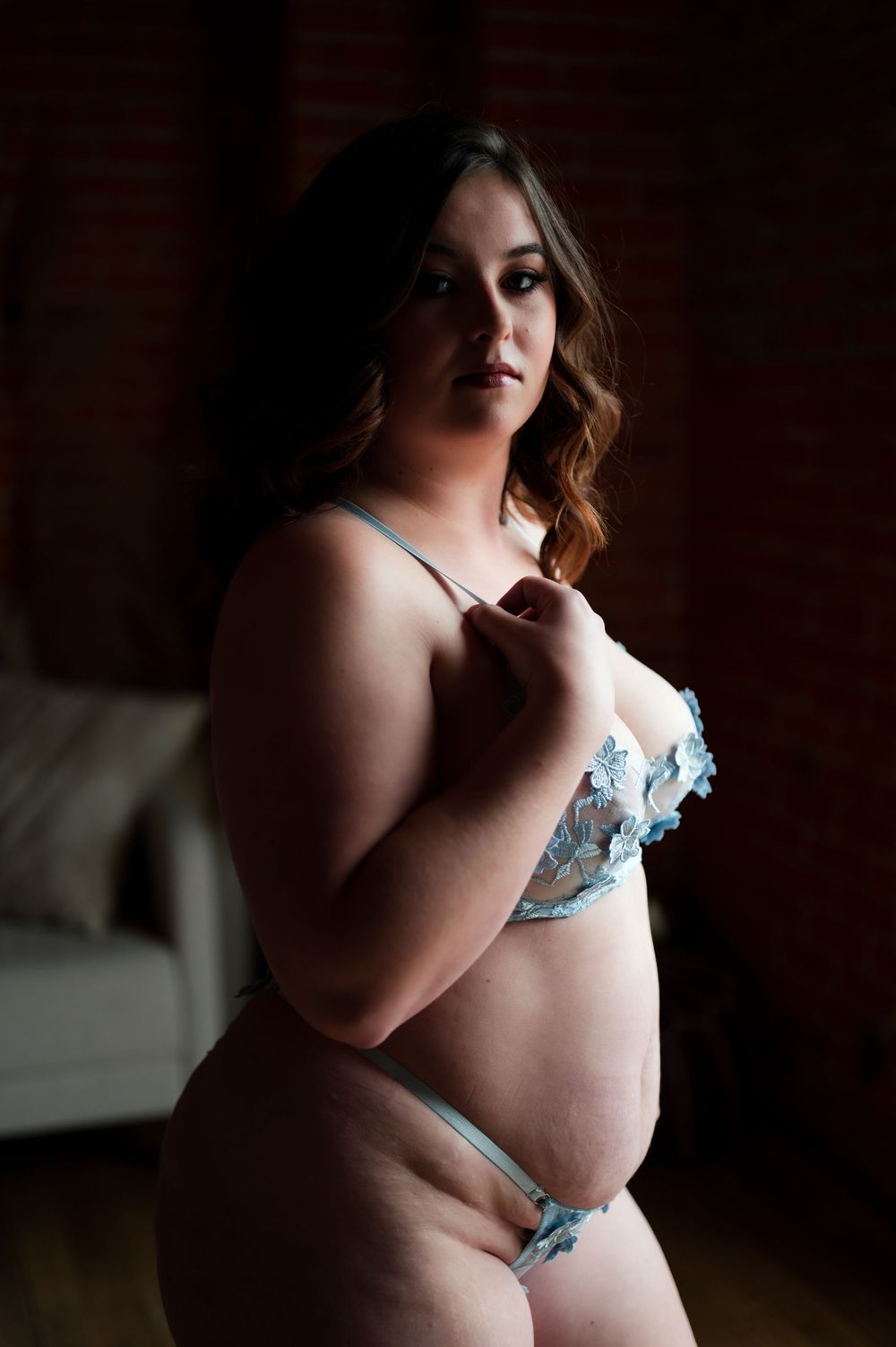 Boudoir photography shot of a person in blue lingerie poses against dark background with dramatic lighting.