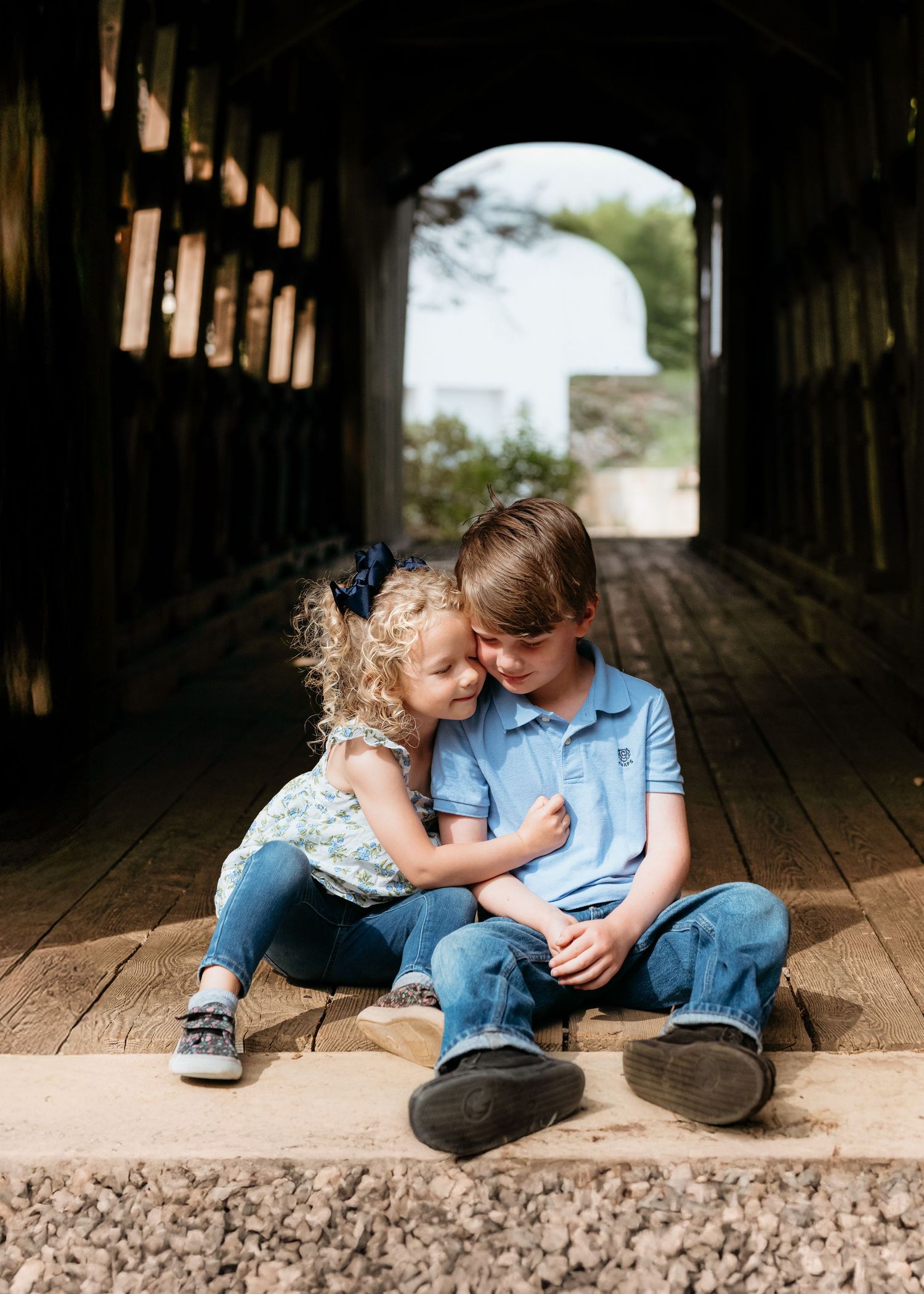Two young siblings share a tender moment sitting on wooden floor inside a covered bridge.