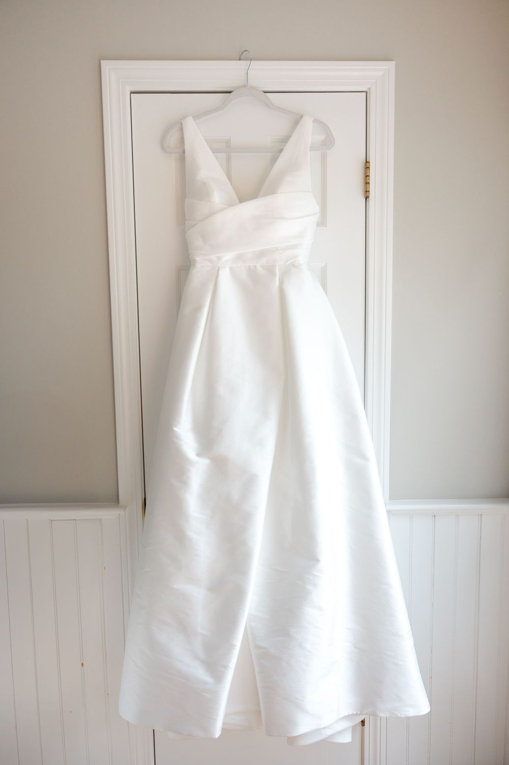 A mikado wedding dress with V-neck design hangs from wooden closet door during the getting ready part of a wedding day in Boston, MA.