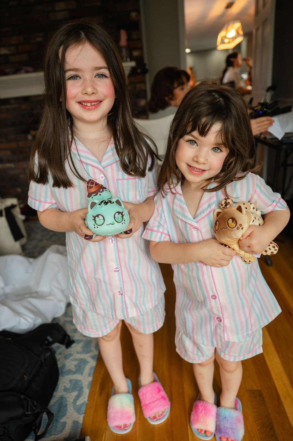 Two children wearing matching pink striped pajamas hold cute plush toys while standing in slippers on hardwood floor.