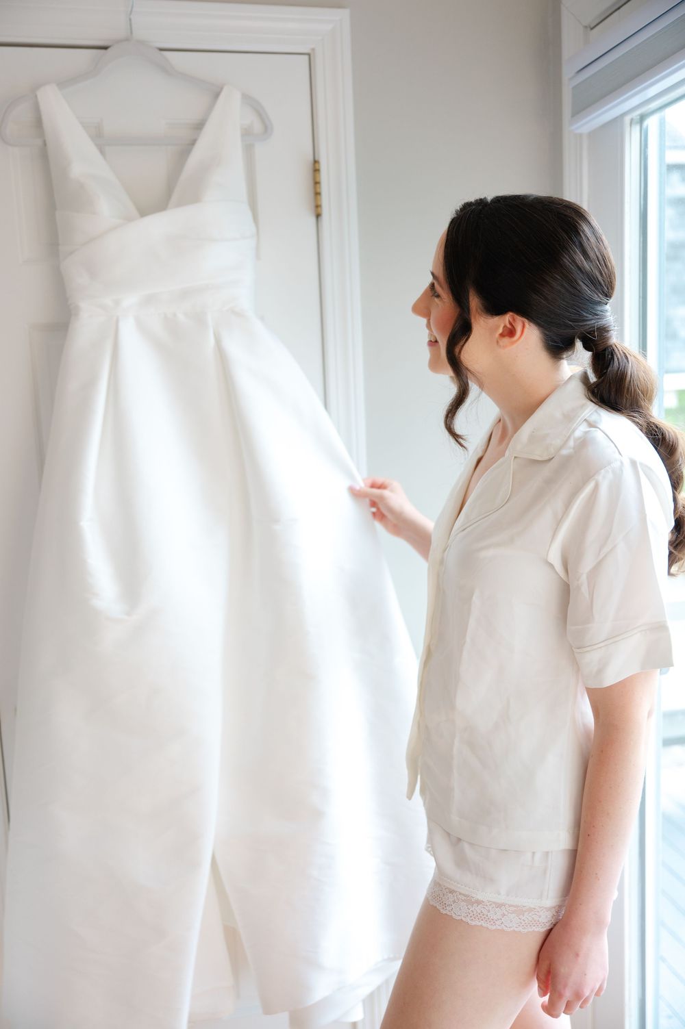 A bride smiles as she looks at her wedding dress  hanging from a closet rod near a window on her wedding morning in Boston, MA