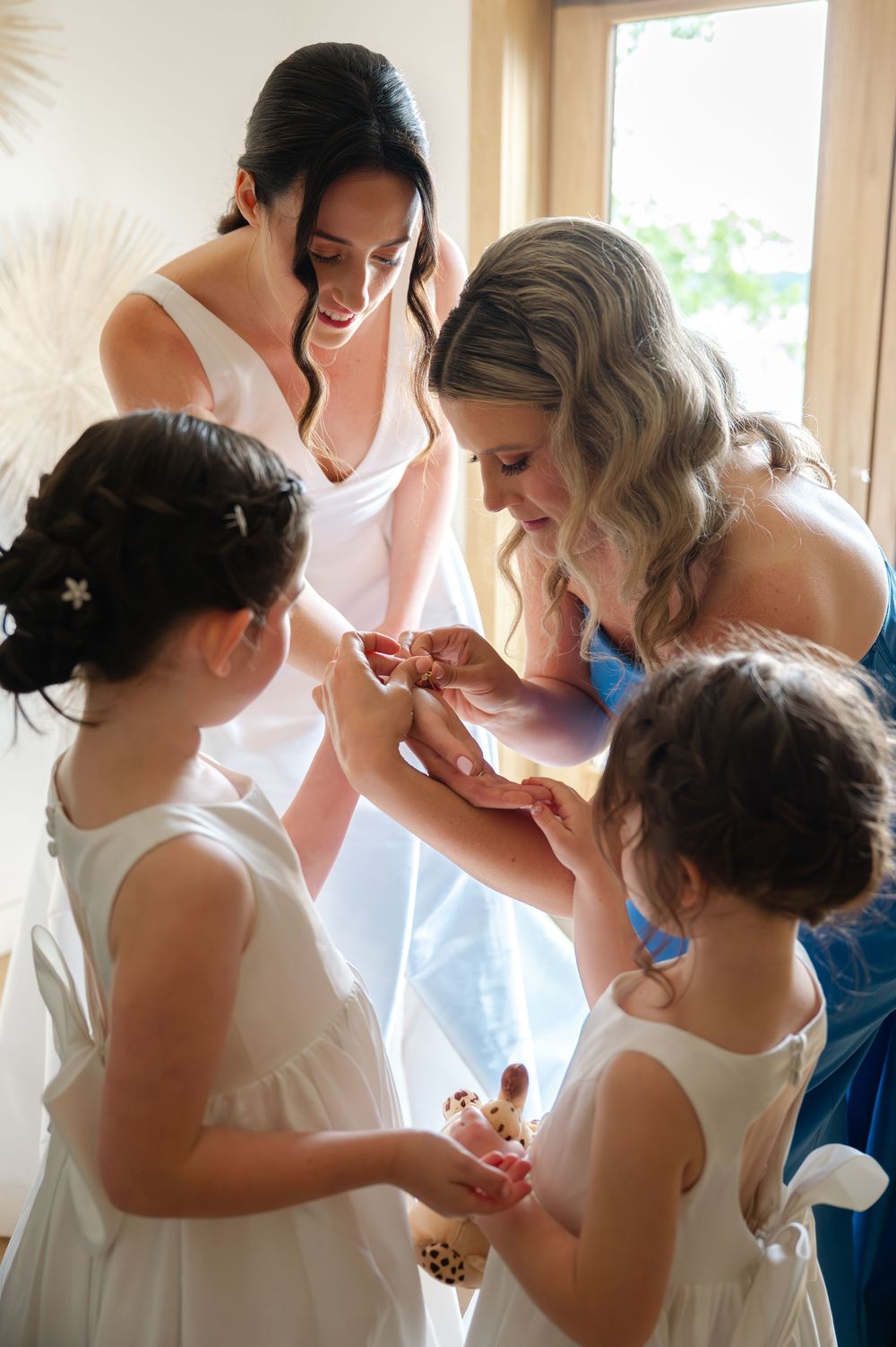 A joyful wedding preparation moment shows a bridesmaid and two flower girls helping the bride put on her jewelry.