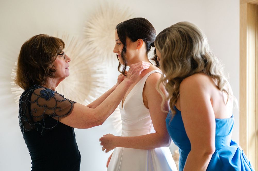 Mother of the bride and maid of honor help with finishing touches to a bride's outfit as she gets ready on her wedding day.