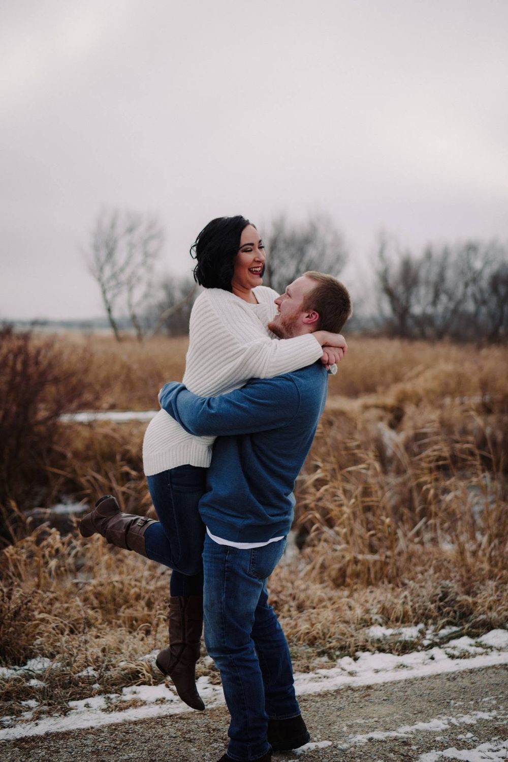 A couple shares a joyful embrace outdoors in winter, man lifting woman in white sweater and jeans near snowy field.