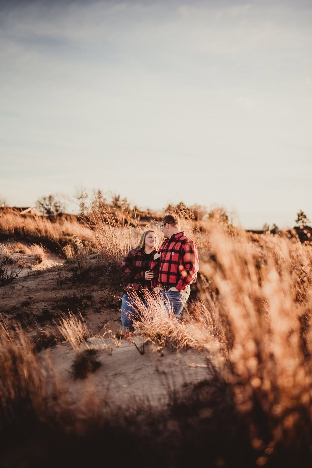 Couple in red plaid jackets embracing among golden grasses and sand dunes during a warm autumn outdoor photoshoot.