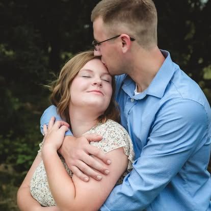 A couple shares a tender embrace outdoors, man in blue shirt kisses womans forehead among lush green trees.