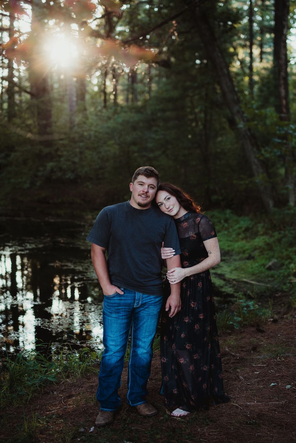 A couple poses together by a serene pond in a wooded setting at sunset.
