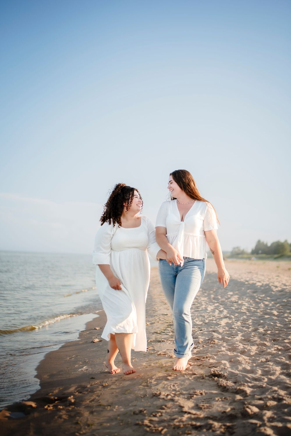 Two people walking barefoot along beach shoreline in casual summer outfits.