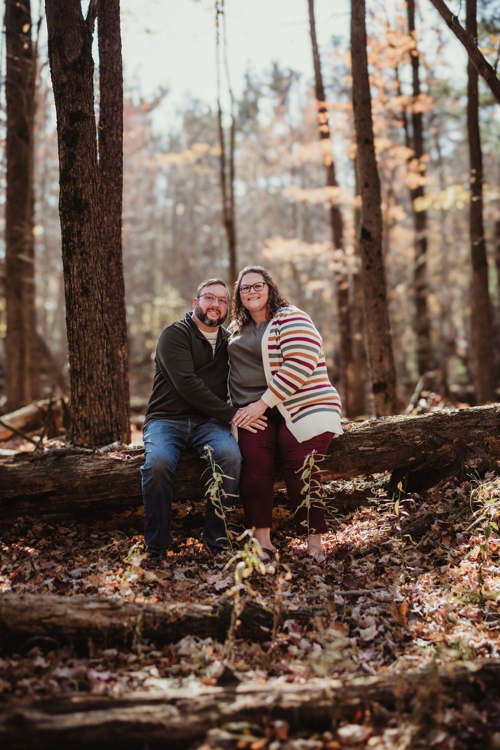 A couple sits together on a fallen log in an autumn forest setting.