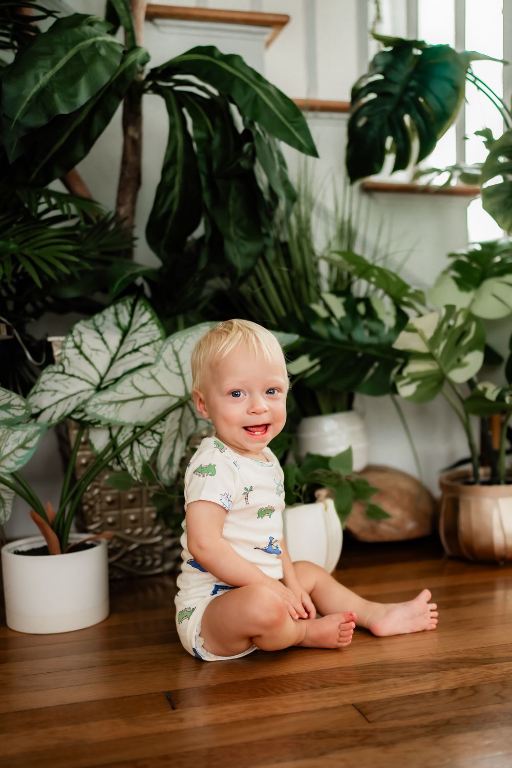 Baby in white onesie sits on wooden table surrounded by lush indoor plants and white planters.