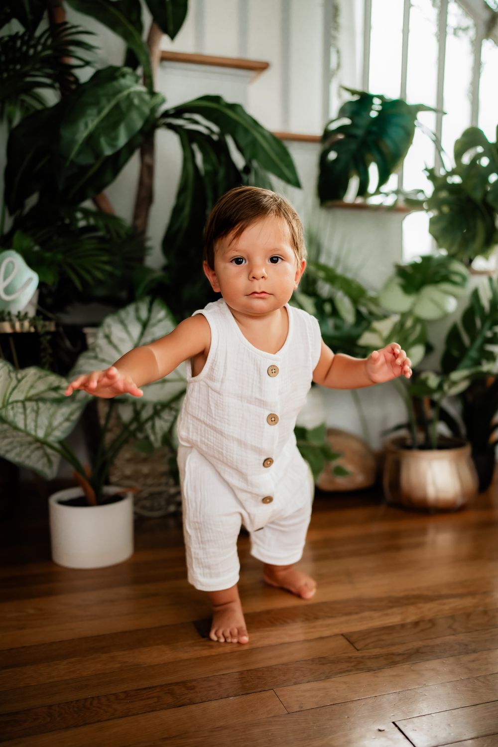 A toddler stands barefoot on hardwood flooring wearing a white button-up romper, surrounded by potted houseplants.