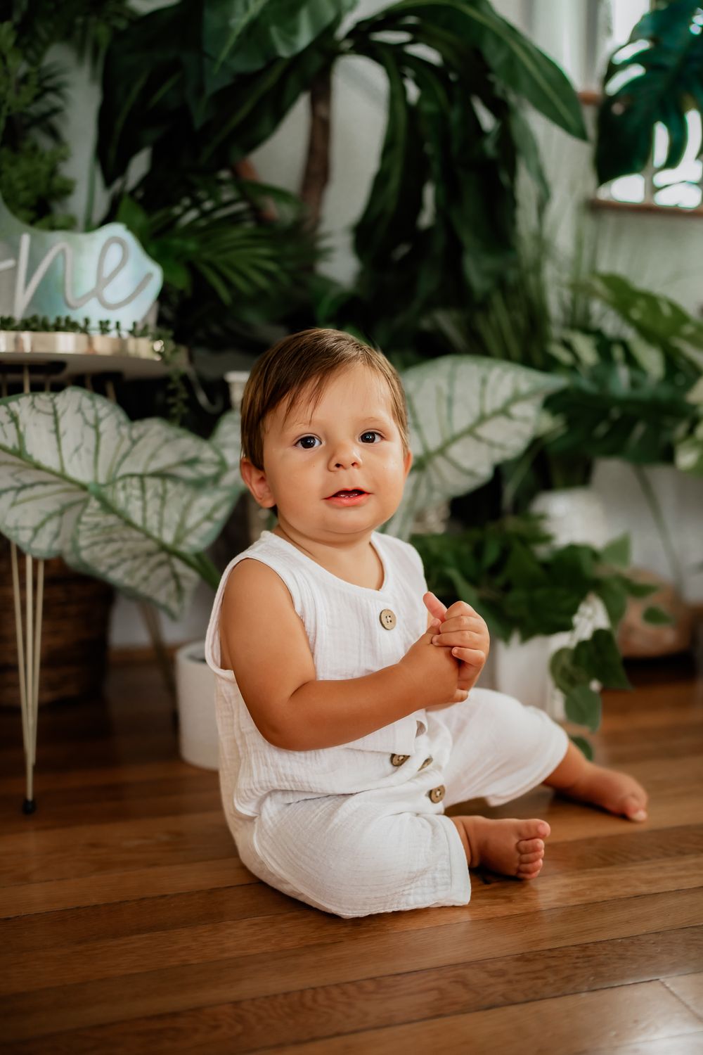Baby in white outfit sitting on wooden floor with lush green plants in background.