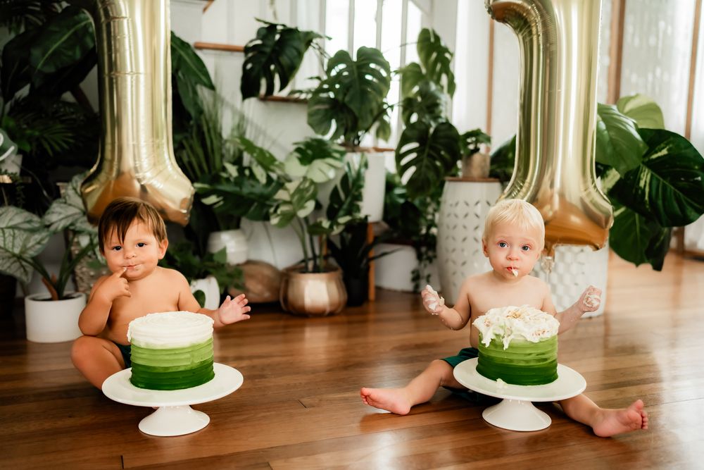 Baby in white attire near birthday cake with green frosting on wooden floor surrounded by plants.