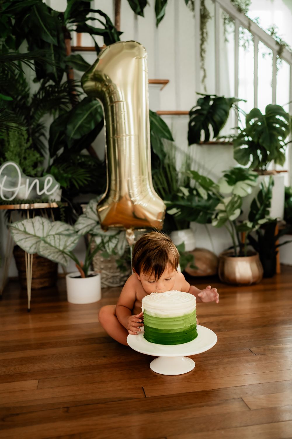 Small green and white cake on white pedestal with large gold balloon and houseplants in background.