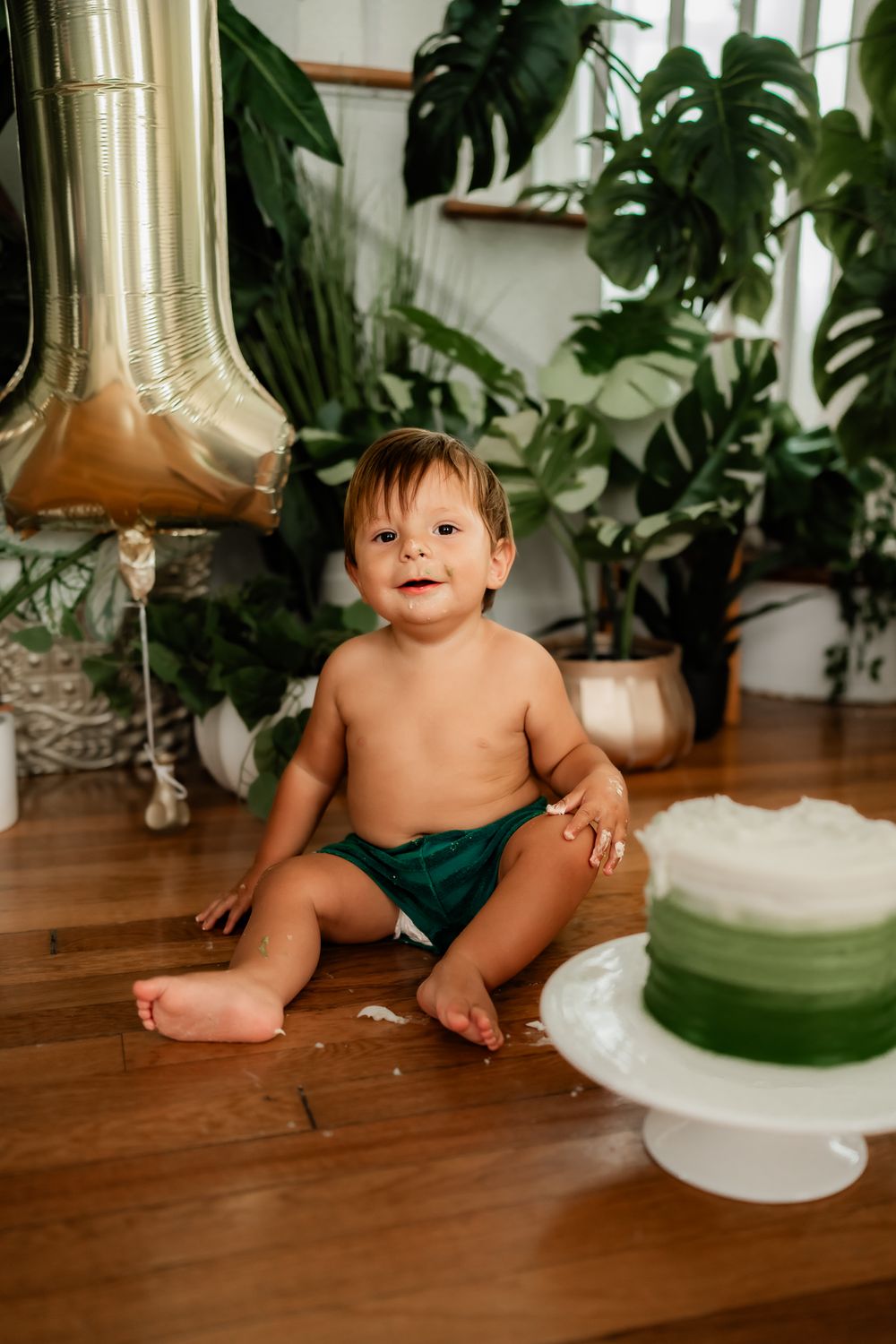 Baby in dark pants sits on hardwood floor surrounded by houseplants and a white birthday cake with balloons.