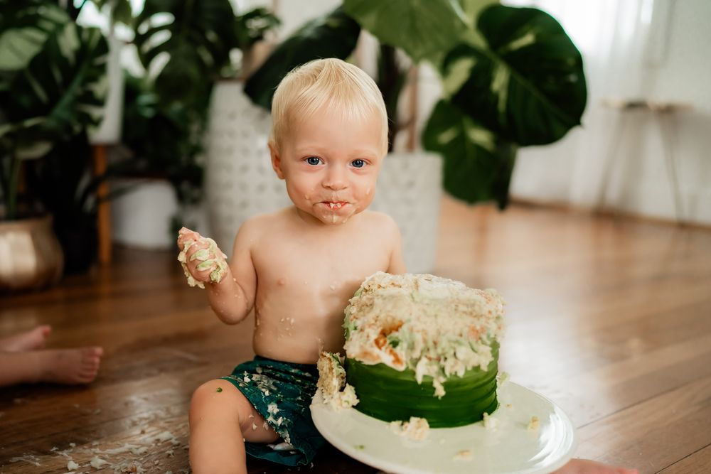 A baby in a highchair enjoying a celebratory cake smash with a green cake on a wooden dining table.