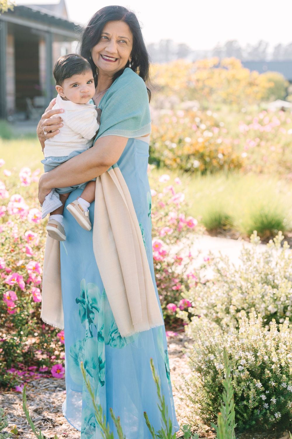 Woman in blue floral dress holding child in garden setting with pink flowers.