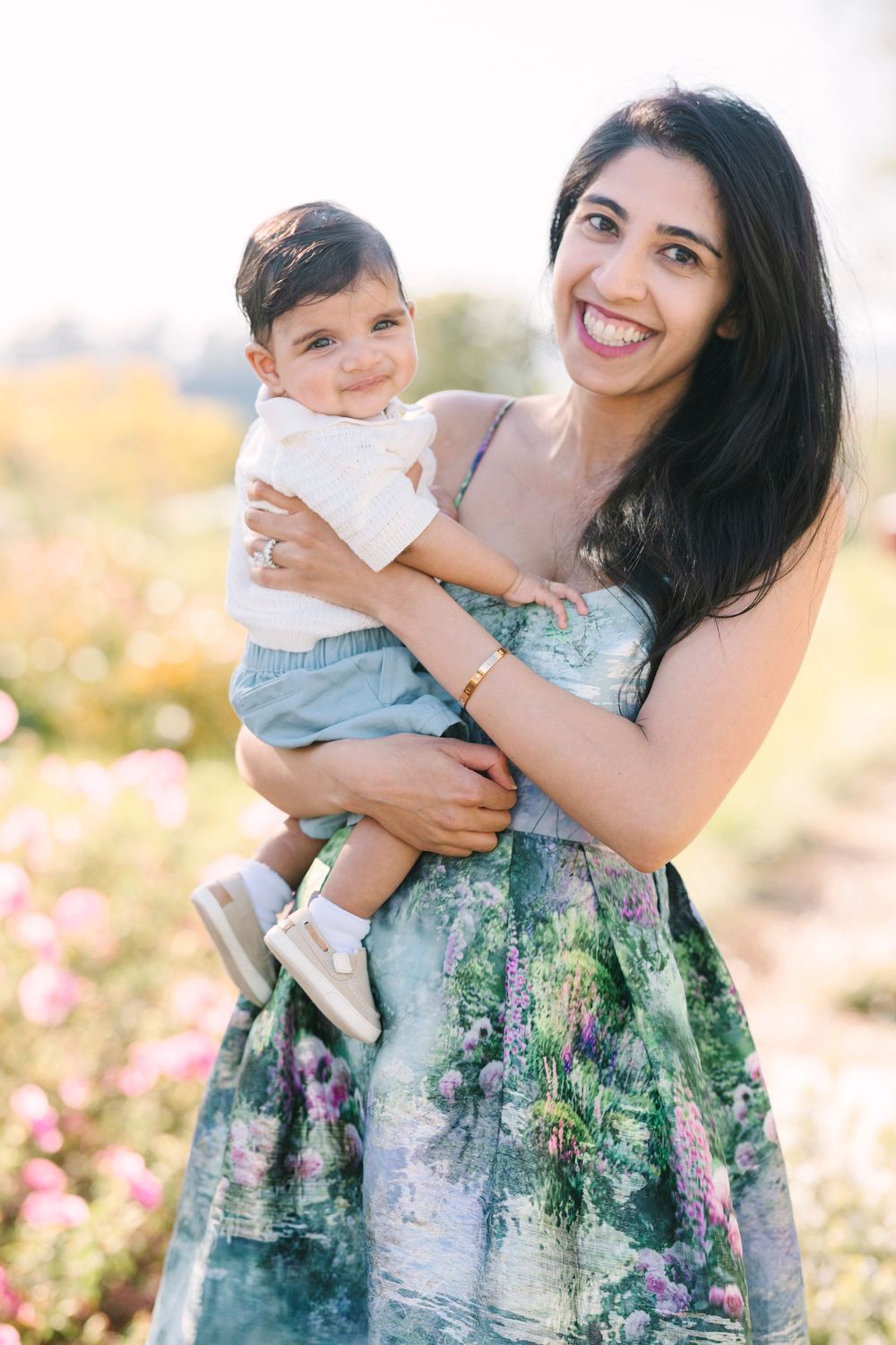Woman in floral dress holding child in sunny garden with pink flowers.