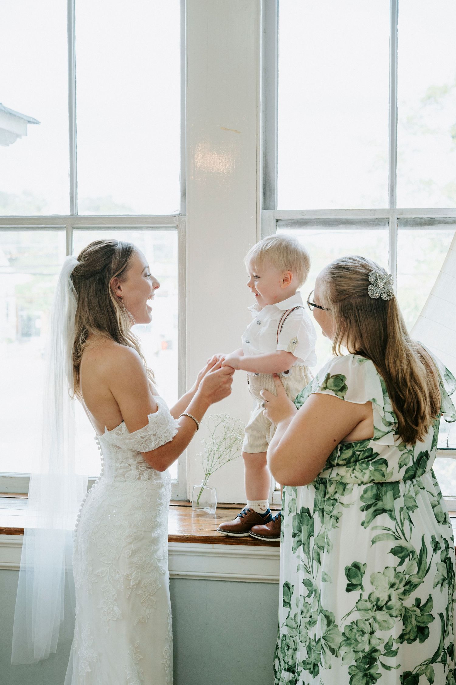 Bridal party dresses in white and floral green patterns interact near bright windows in a candid moment.