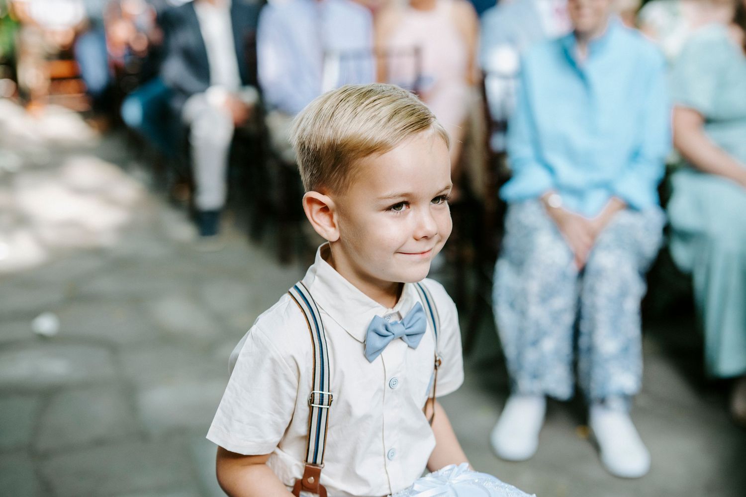 A young student in a white shirt and suspenders stands outside with a blurred background on what appears to be a school day.