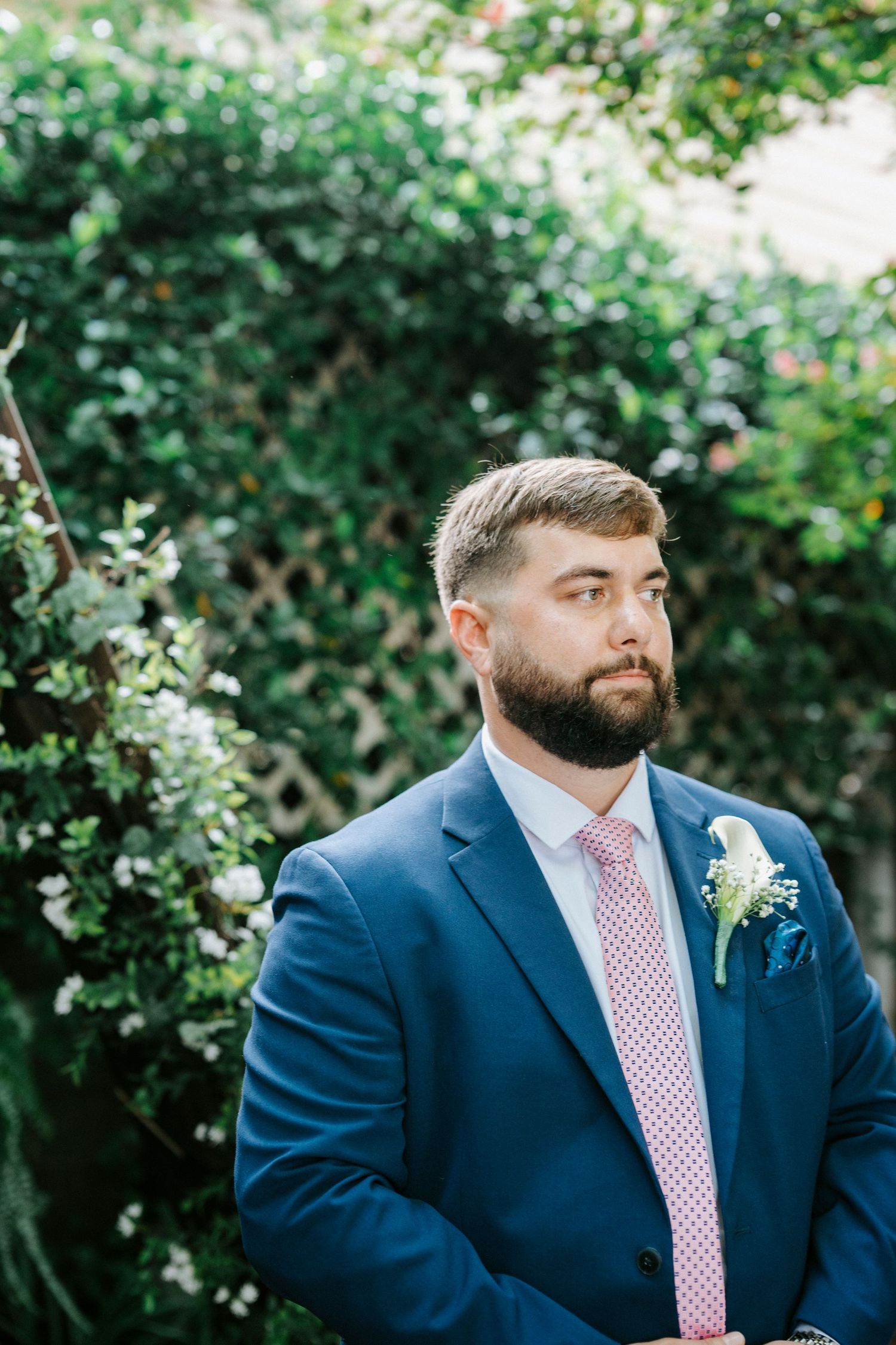 Groom in a navy suit with pink striped tie stands in a lush garden setting.