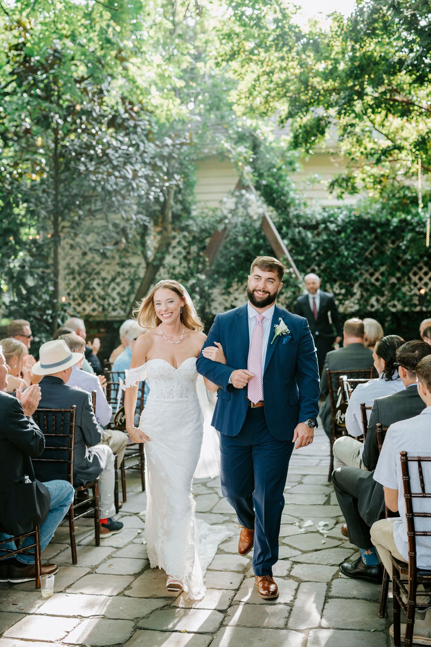Wedding guests seated outdoors watch as a couple makes their recessional exit down a stone path after the ceremony.