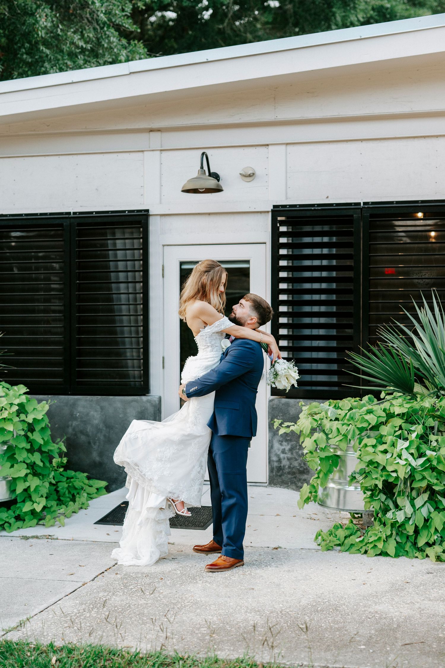 Wedding couple embraces and kisses in front of a white building with black shutters.