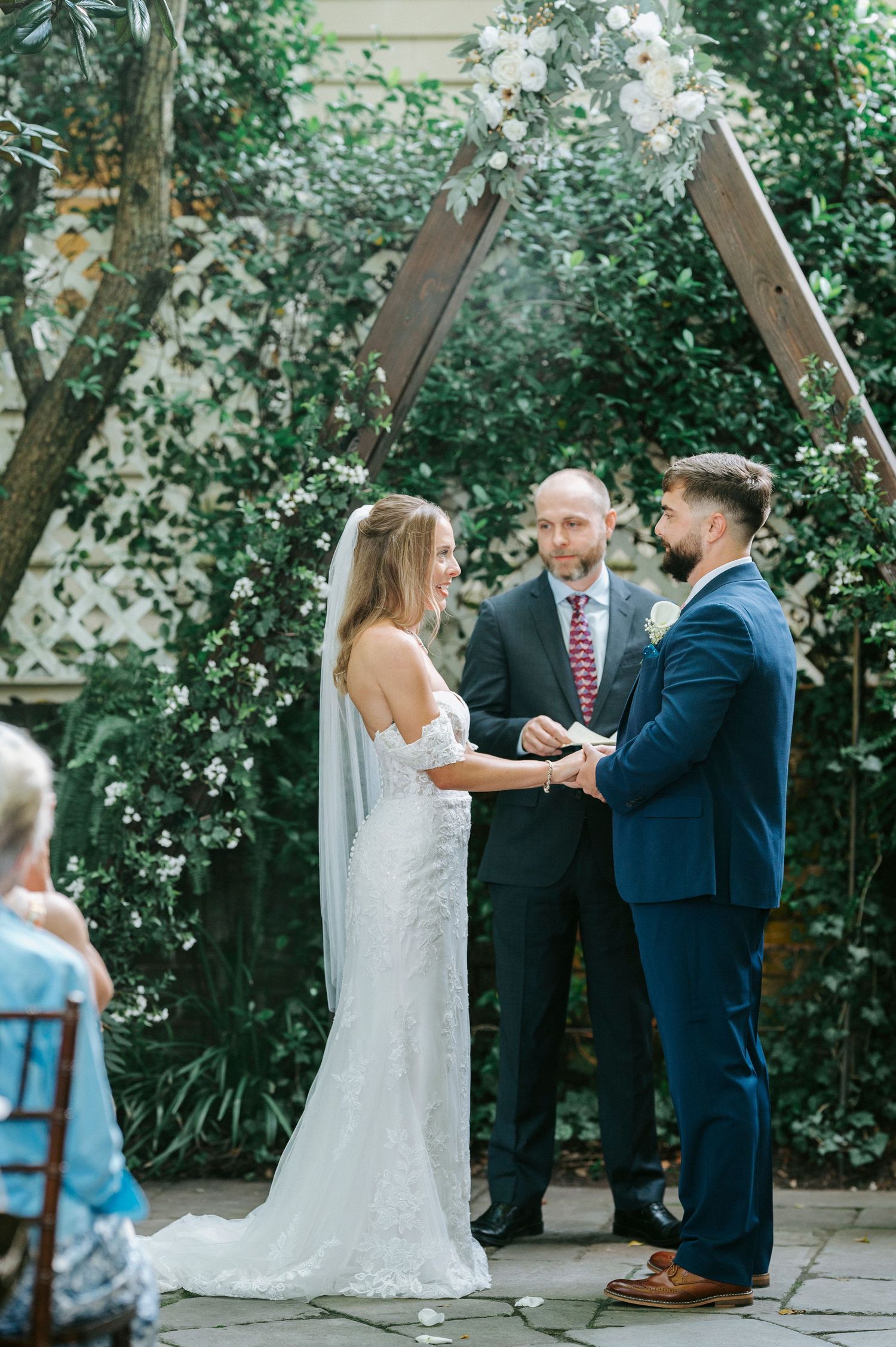 Wedding ceremony takes place in a garden setting with white flowers and greenery.