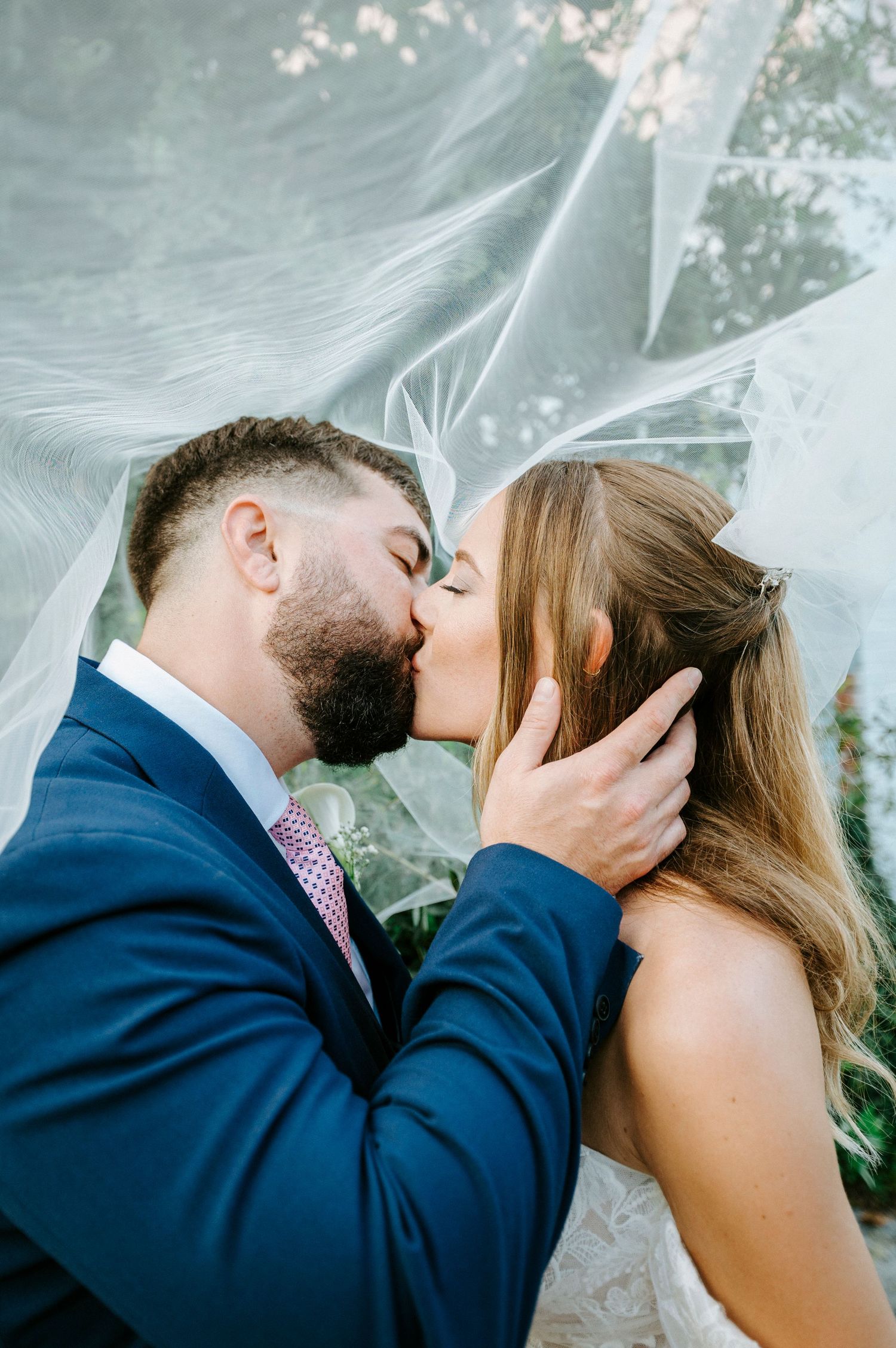 Romantic couple share a tender kiss under a flowing white veil at an outdoor wedding ceremony.