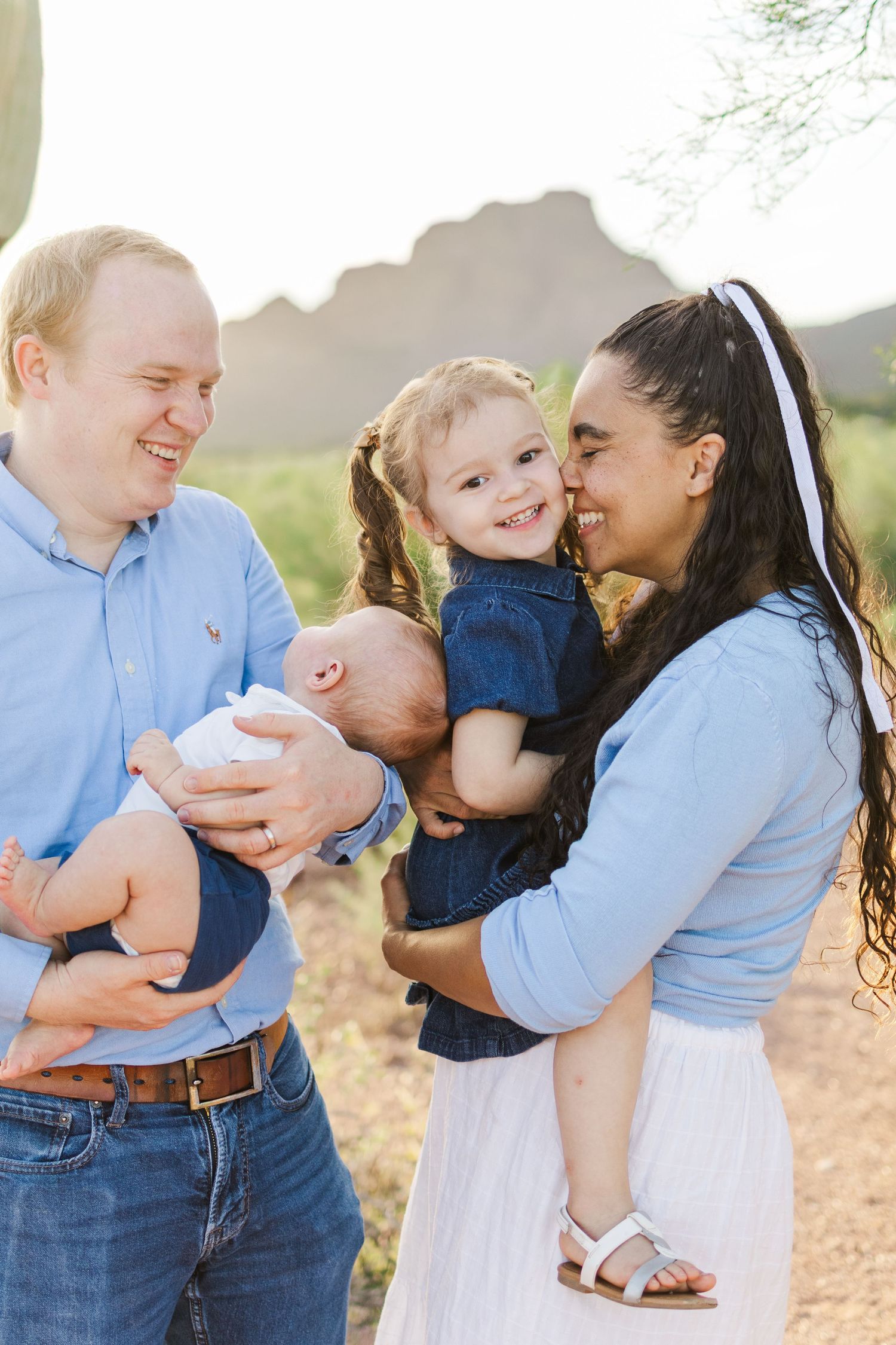 Young family of 4 in the desert with a mountain in the background as mom tickles the toddler girls cheek with her nose and dad holds infant.