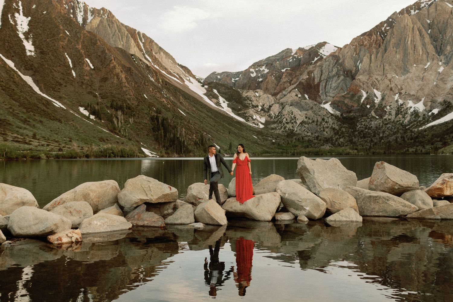 Couple in red dress and suit standing on rocks by a mountain lake with reflection.