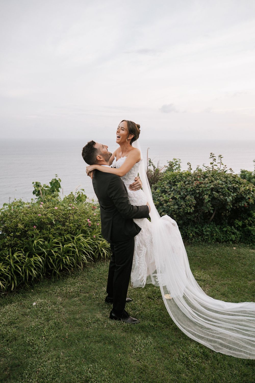 A romantic wedding couple embraces on a coastal clifftop with ocean views and lush greenery in the background.