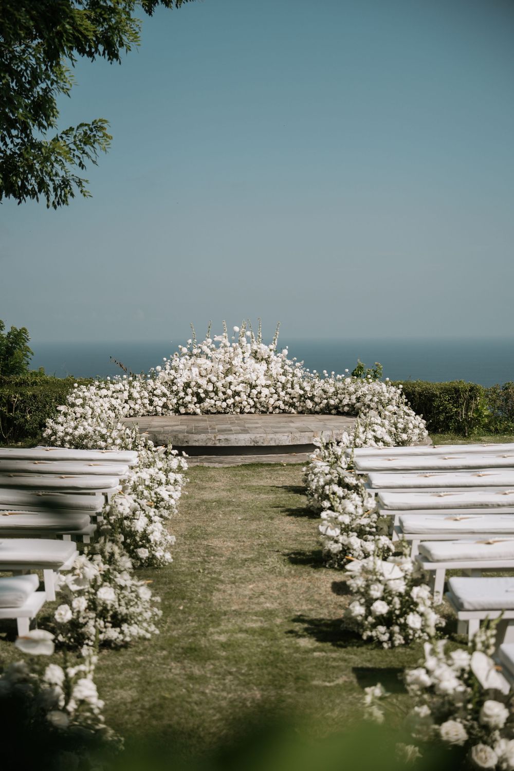 White floral arch arrangements line outdoor wooden benches at an elegant oceanfront wedding ceremony setup.