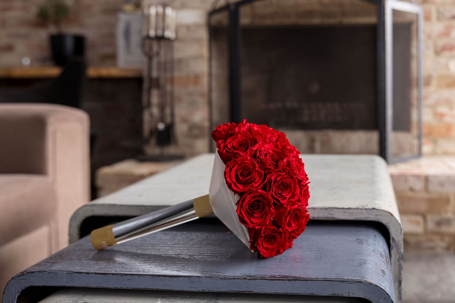 A wedding floral bouquet of red roses sits on a table by a fire place.