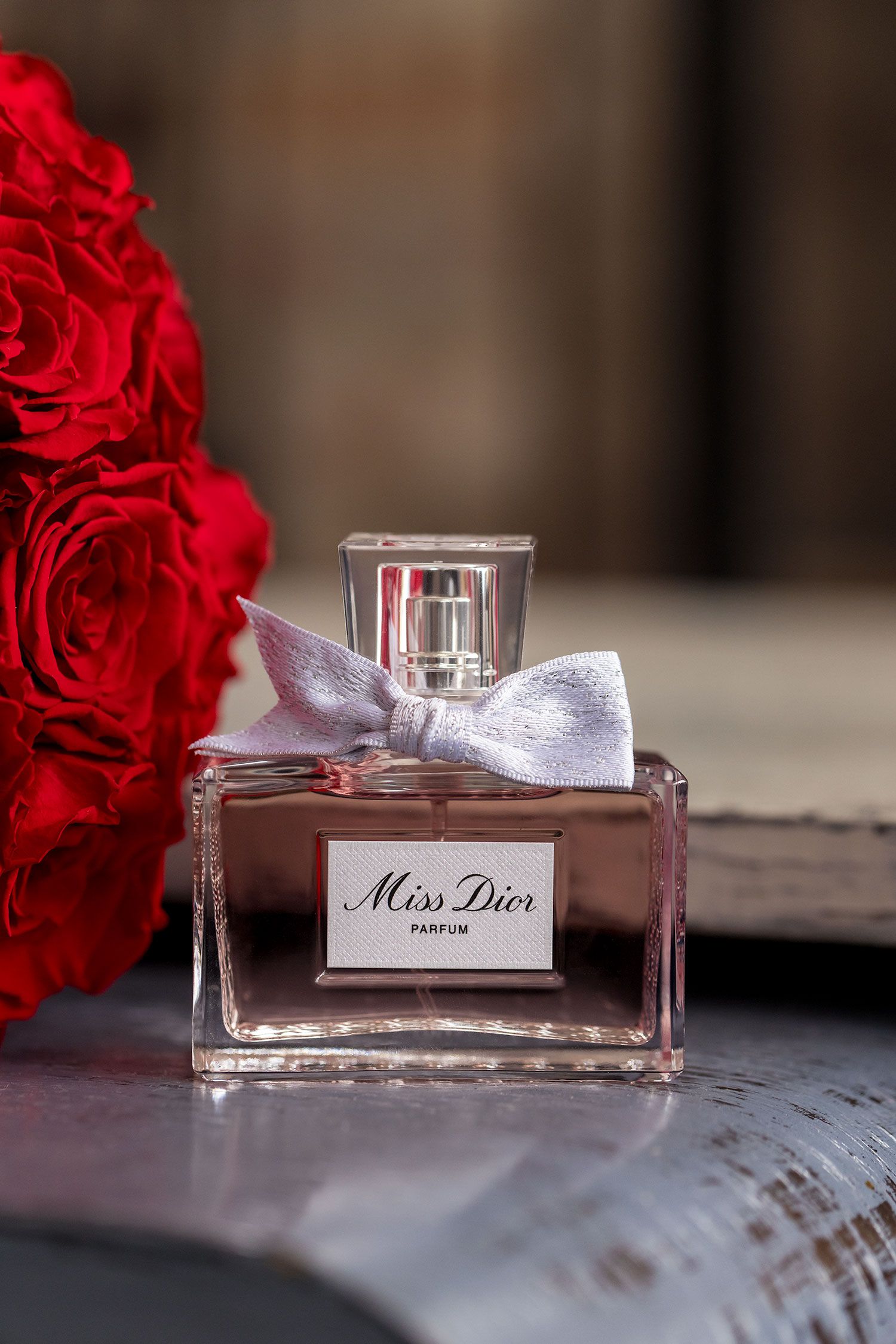 A bottle of Miss Dior perfume sits elegantly beside vibrant red roses on a rustic wooden table.