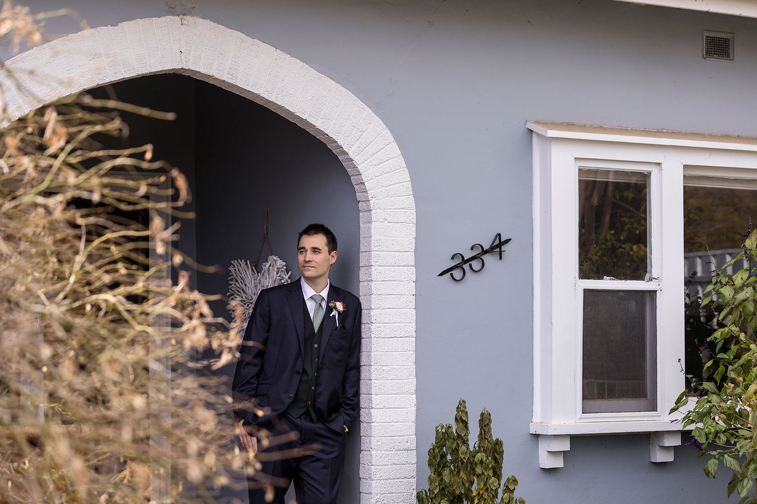 Groom standing in the Archway entrance of an old  House during wedding Preparations wearing his Oxford wedding suit with button hole flower.