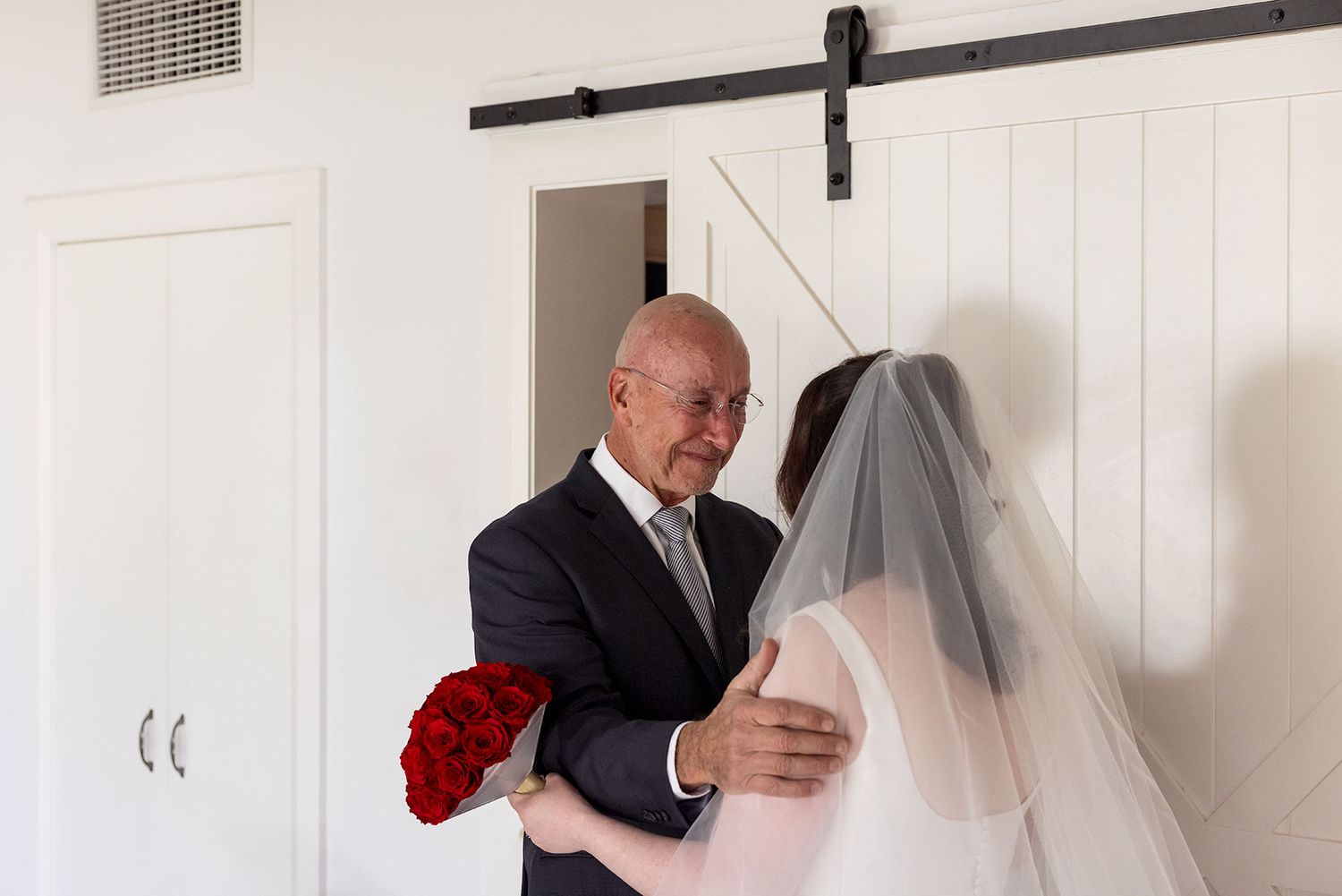 A bride in a white veil and dress stands with her Dad in a dark suit, surrounded by white walls, seeing her for the first time in her dress.